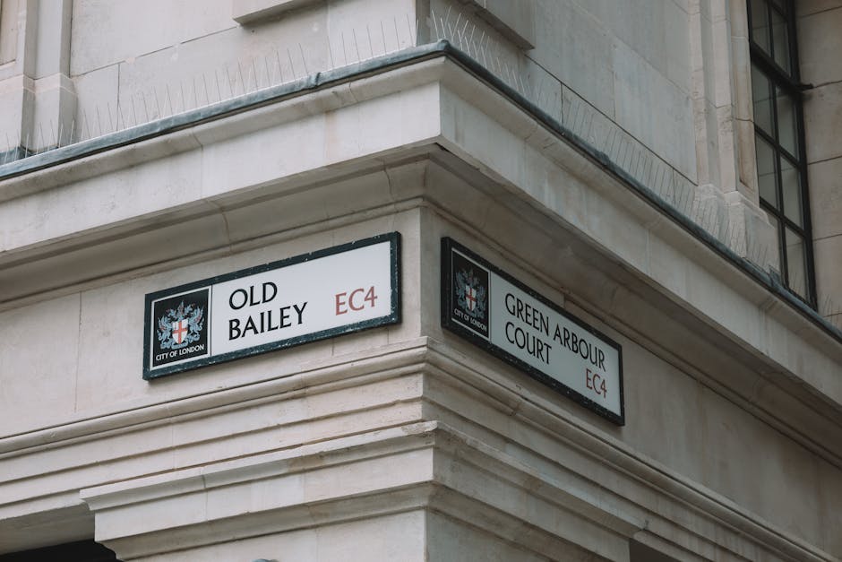 A close-up view of the corner of a beige stone building in London, featuring two street signs attached to the facade. The left sign reads 'OLD BAILEY EC4' with the City of London crest above, and the