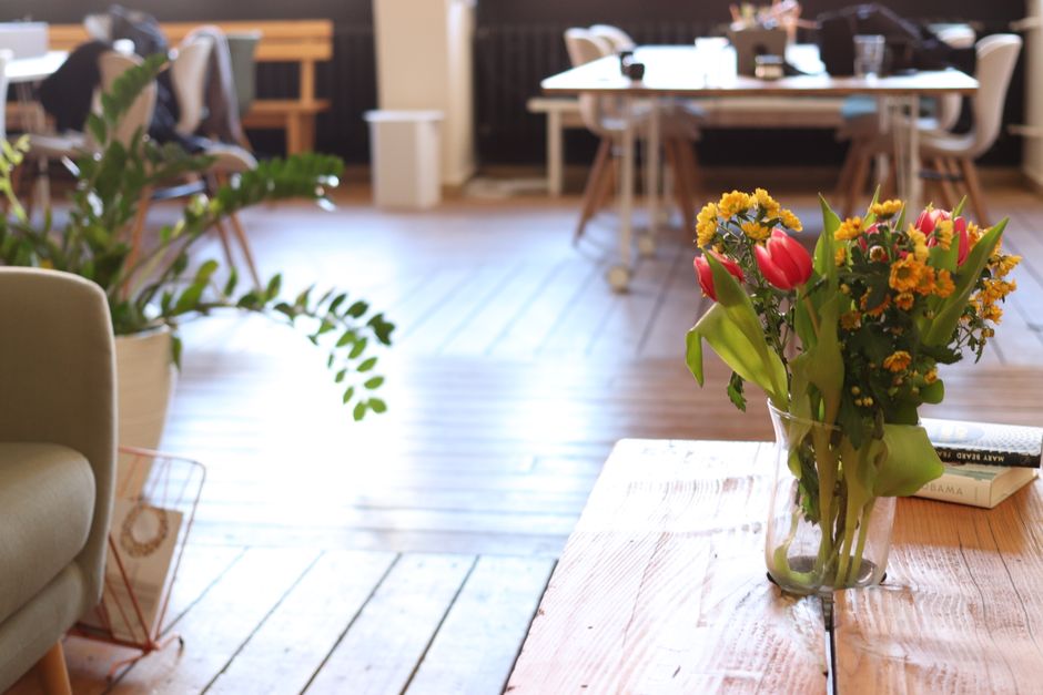A clear glass vase filled with a fresh floral bouquet featuring red tulips, small yellow daisies, and green foliage, placed on a light wooden table inside a bright, modern office or cafe setting. The
