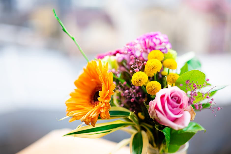 A vibrant floral arrangement featuring a bright orange gerbera daisy, a soft pink rose, clusters of small yellow billy balls, and pink hypericum berries, all set in a mixed bouquet. The flowers are wr