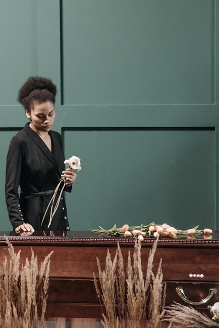 A young woman with dark, curly hair styled in a natural afro, dressed in a black blazer, stands beside a wooden casket in a dimly lit room with muted green walls. She holds a small, white flower and l