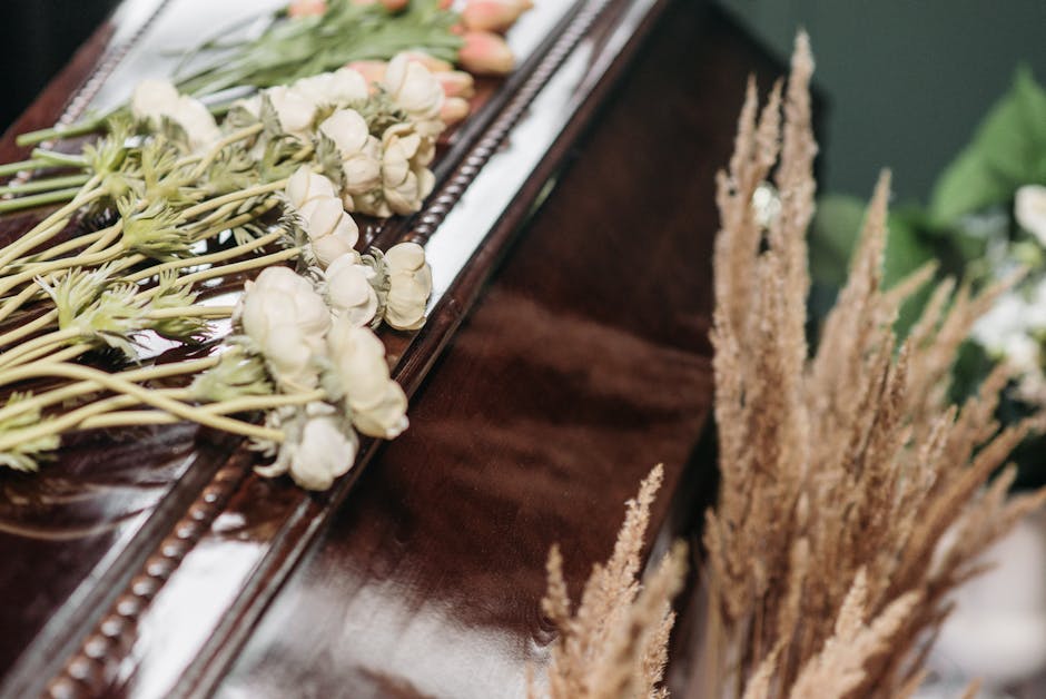 Close-up of a floral arrangement featuring white roses with delicate petals, arranged on a dark polished wooden surface, possibly part of a funeral floral tribute. The white roses are positioned in a