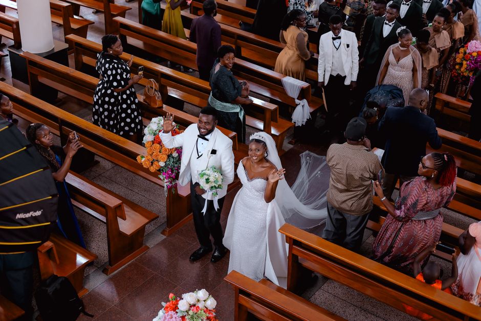 A newlywed couple is walking down the aisle inside a church, with the groom in a white tuxedo and the bride in a white wedding gown, both smiling and waving at guests. The bride is wearing a veil and