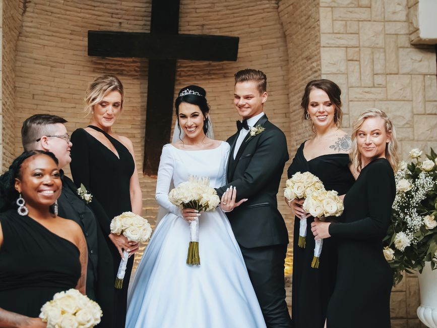 A bride and groom stand centrally inside a church with a brick wall and a large wooden cross behind them, surrounded by five bridesmaids and a groomsman. The bride wears a long-sleeved white wedding g